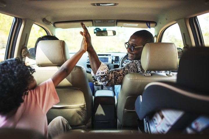 Black Dad in front seat of car high-fiving his daughter in the backseat - holiday season road trip