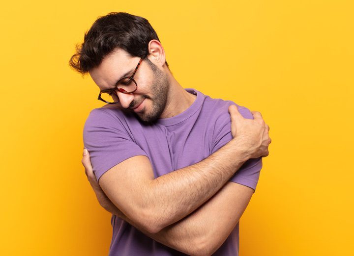 handsome man in glasses and a purple t-shirt hugging himself against a yellow background - self-love
